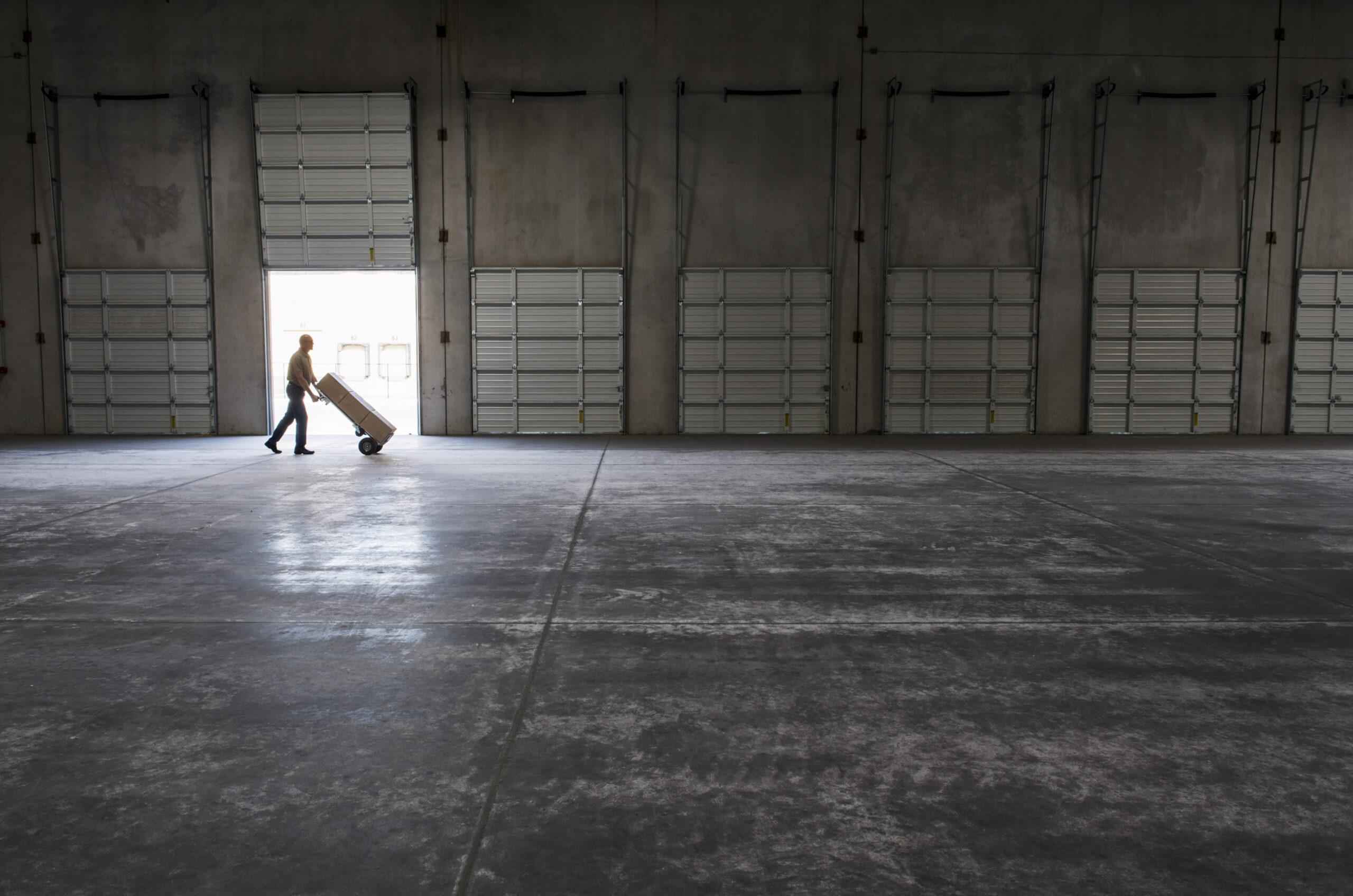 Man walking with loaded hand truck past loading dock doors in new empty warehouse. Concrete Warehouse