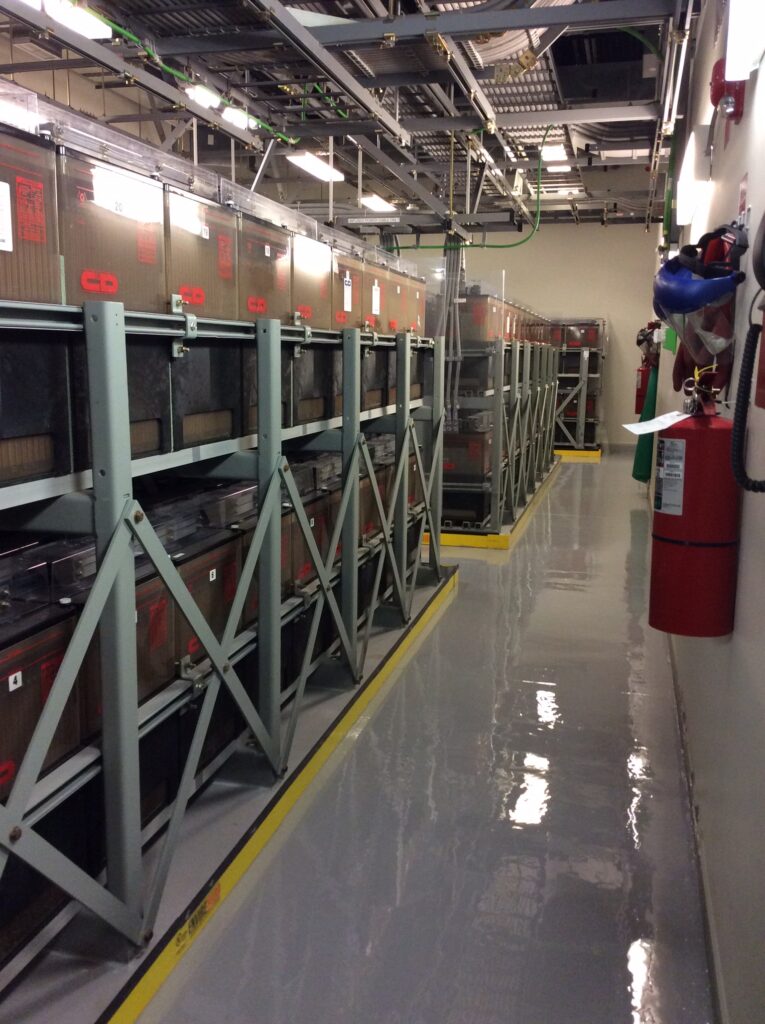 Glossy gray coated floor in a battery room aisle with racks of backup batteries and overhead cable trays.